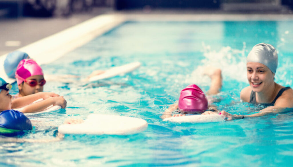 Children learning to swim in a pool with an instructor. Four kids in colorful swim caps hold onto kickboards while practicing basic swimming techniques. A smiling swim coach in a silver cap guides and encourages them in the water. This image highlights a group swim lesson, showcasing an organized and engaging class environment scheduled by swim lesson software.