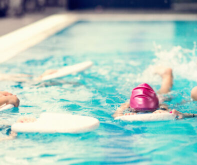 Children learning to swim in a pool with an instructor. Four kids in colorful swim caps hold onto kickboards while practicing basic swimming techniques. A smiling swim coach in a silver cap guides and encourages them in the water. This image highlights a group swim lesson, showcasing an organized and engaging class environment scheduled by swim lesson software.