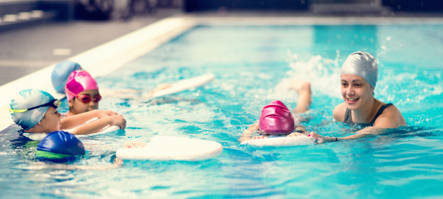 Children learning to swim in a pool with an instructor. Four kids in colorful swim caps hold onto kickboards while practicing basic swimming techniques. A smiling swim coach in a silver cap guides and encourages them in the water. This image highlights a group swim lesson, showcasing an organized and engaging class environment scheduled by swim lesson software.