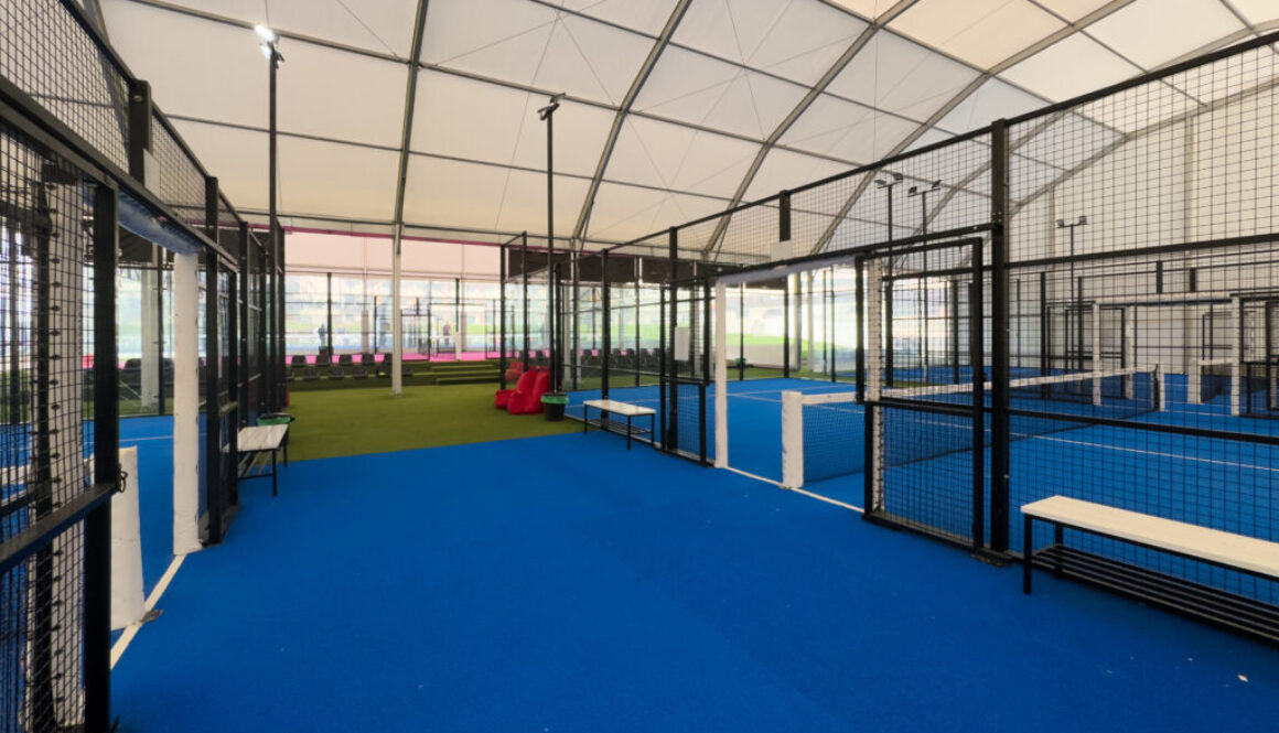 Spacious indoor padel court featuring a vivid blue surface, surrounded by metal fences and overhead bright lights, perfect for training or competitive matches within a well-maintained sports facility.