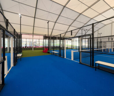 Spacious indoor padel court featuring a vivid blue surface, surrounded by metal fences and overhead bright lights, perfect for training or competitive matches within a well-maintained sports facility.