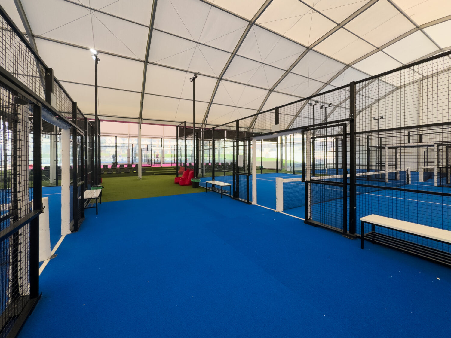 Spacious indoor padel court featuring a vivid blue surface, surrounded by metal fences and overhead bright lights, perfect for training or competitive matches within a well-maintained sports facility.